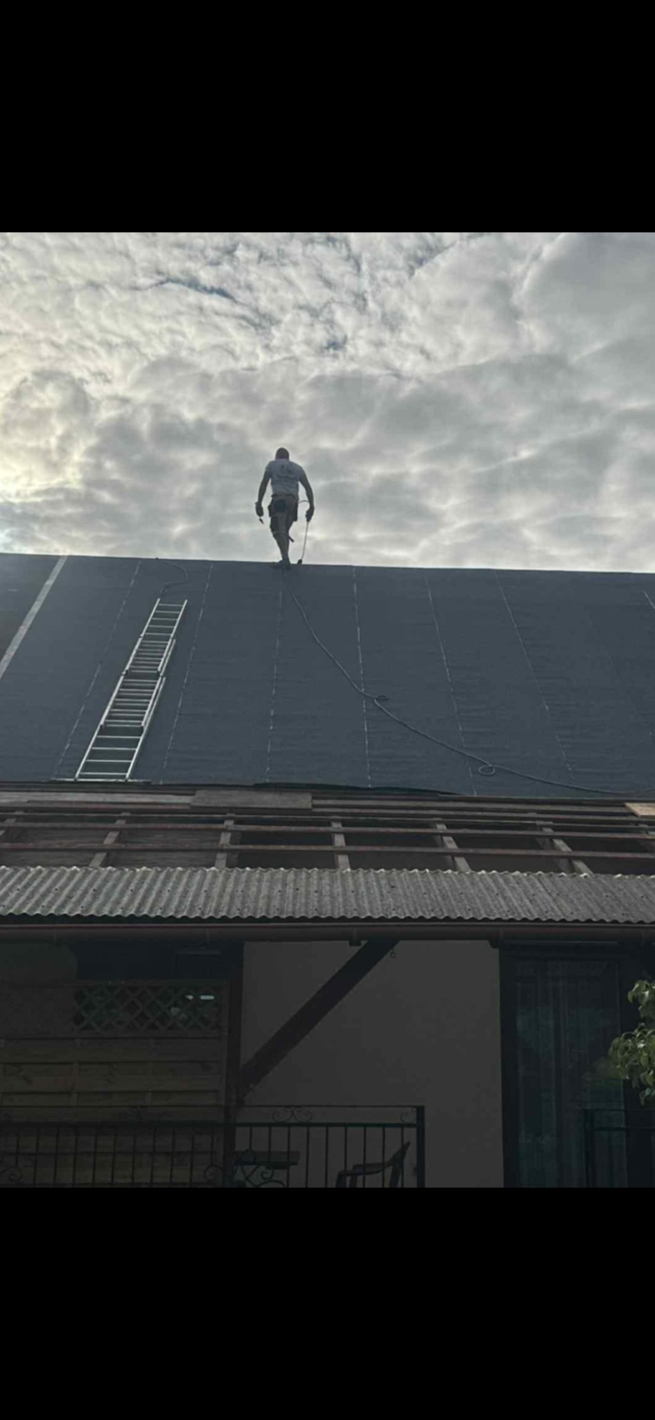 Dekarz na dachu pokrytym papą, drabina oparta o ścianę. Widoczne krokwie i fragment budynku. Pochmurne niebo w tle.