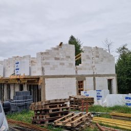 Reflex - Wznoszenie ścian z bloczków Solbet na budowie domu. Widoczne drewniane elementy konstrukcyjne, palety i materiały budowlane. Szary bus na pierwszym planie, zieleń w tle.