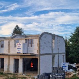 Reflex - Surowy stan dwukondygnacyjnego domu z betonu komórkowego Solbet, w trakcie budowy. Widoczne palety z bloczkami Solbet, tło: drzewa i błękitne niebo.