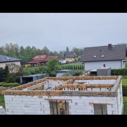 Reflex - Wznoszenie ścian domu z bloczków betonu komórkowego Ytong, z widocznym szalunkiem pod wieniec. W tle zabudowa jednorodzinna, wiosenny krajobraz i pochmurne niebo.