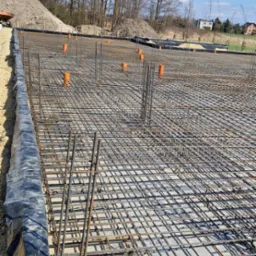 Zbrojenie fundamentów z wystającymi rurami kanalizacyjnymi na tle błękitnego nieba z chmurami i drzewami. Widok z góry na dużą powierzchnię zbrojenia.