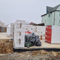 Reflex - Budowa domu w toku: białe bloczki Termobet tworzą ściany na fundamencie. W tle zielony budynek i fragment samochodu dostawczego z naklejką pająka.