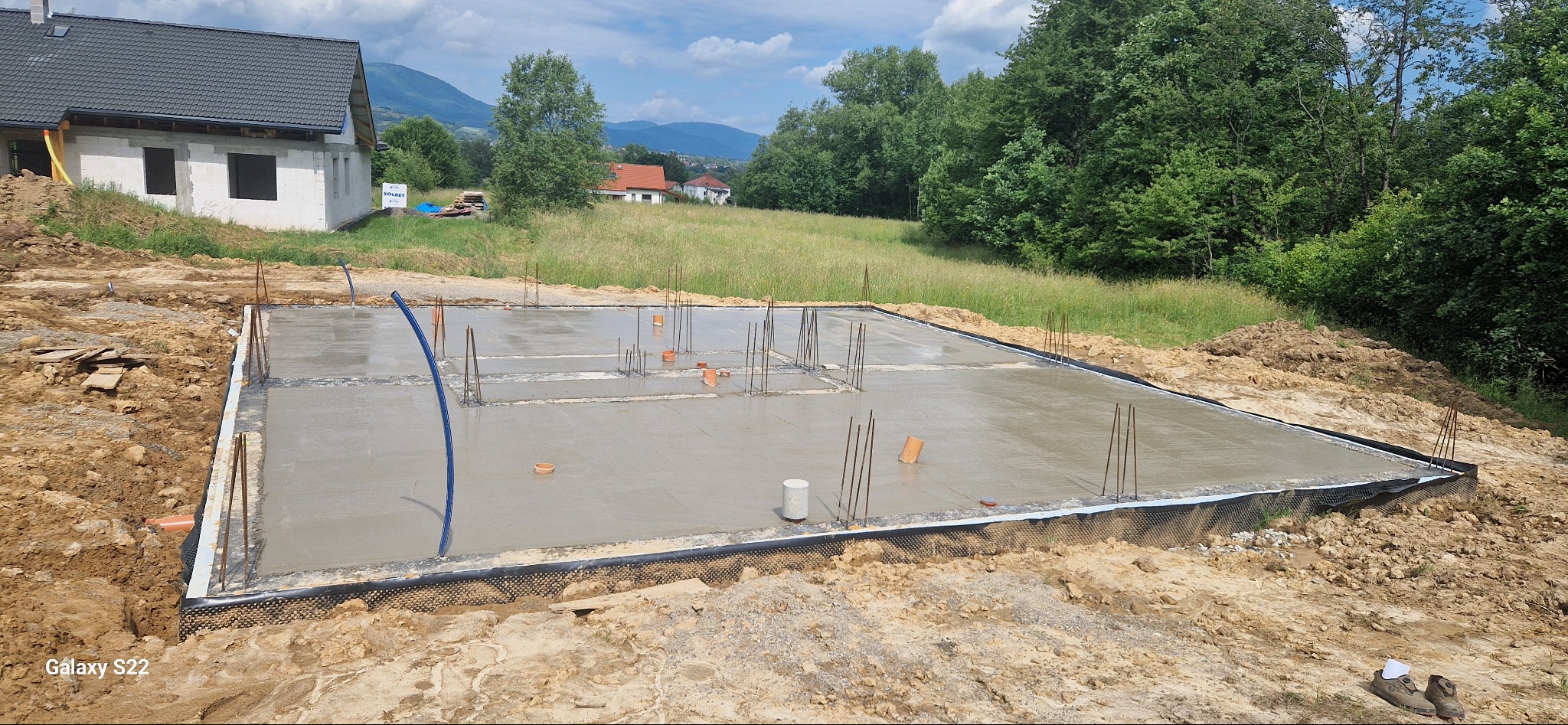 Zalany fundament z wystającymi prętami zbrojeniowymi na tle budującego się domu i zieleni. Widok na świeży beton, elementy instalacji i hydroizolację.