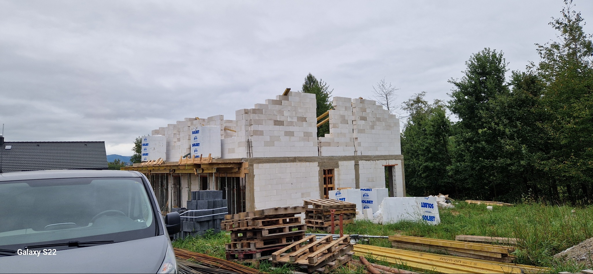 Wznoszenie ścian z bloczków Solbet na budowie domu. Widoczne drewniane elementy konstrukcyjne, palety i materiały budowlane. Szary bus na pierwszym planie, zieleń w tle.