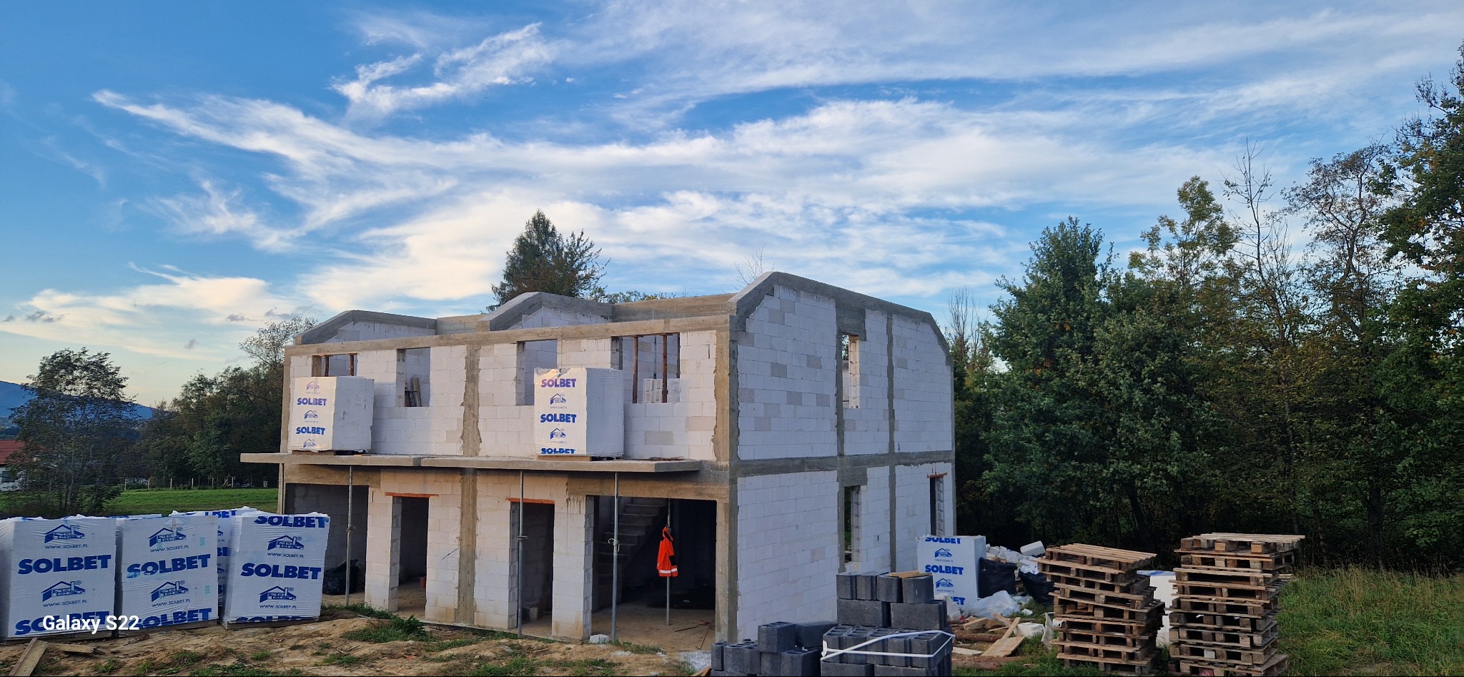 Surowy stan dwukondygnacyjnego domu z betonu komórkowego Solbet, w trakcie budowy. Widoczne palety z bloczkami Solbet, tło: drzewa i błękitne niebo.