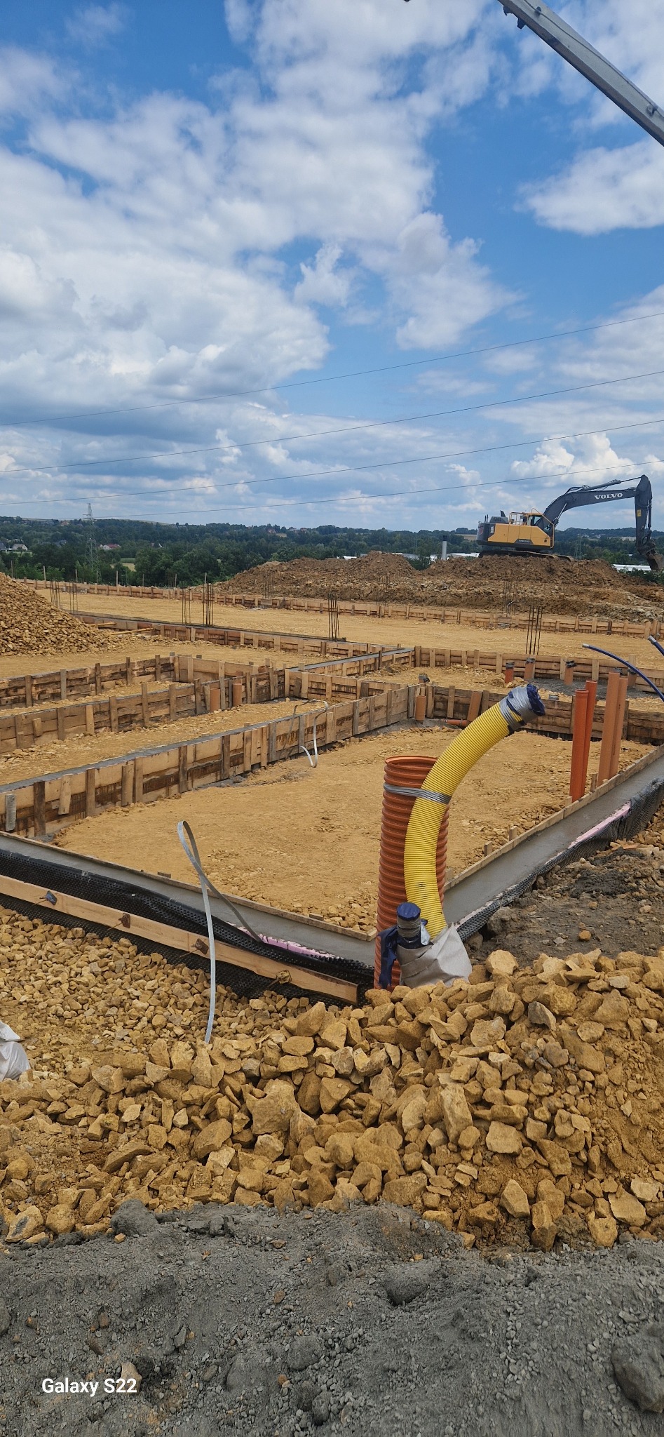 Przygotowanie terenu pod fundamenty z użyciem szalunków drewnianych, rur drenażowych i ciężkiego sprzętu budowlanego Volvo w tle, na tle błękitnego nieba.