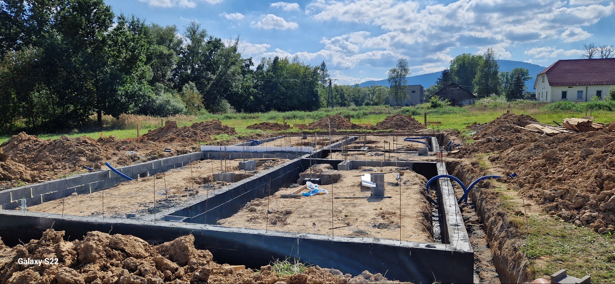 Zalane fundamenty domu jednorodzinnego z wystającymi prętami zbrojeniowymi, otoczone ziemią i z widokiem na zielone pola oraz budynki w tle.