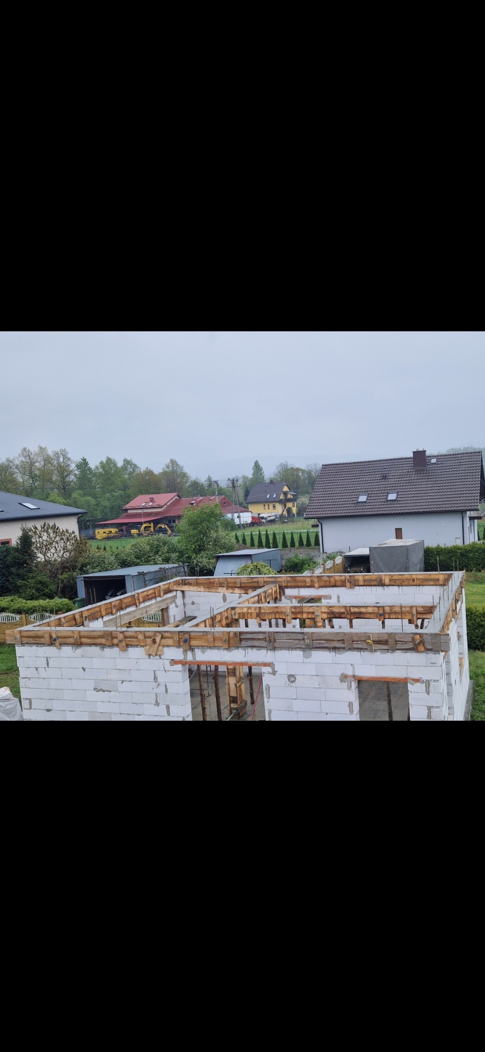Wznoszenie ścian domu z bloczków betonu komórkowego Ytong, z widocznym szalunkiem pod wieniec. W tle zabudowa jednorodzinna, wiosenny krajobraz i pochmurne niebo.