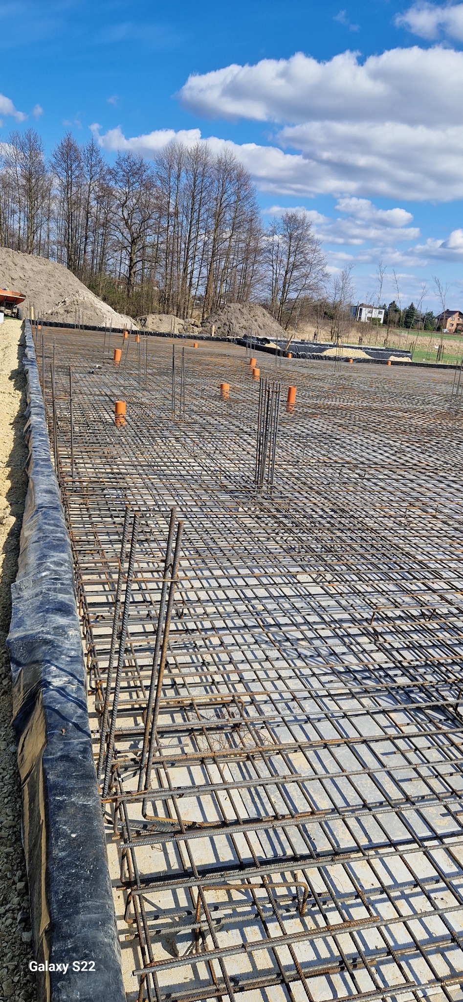 Zbrojenie fundamentów z wystającymi rurami kanalizacyjnymi na tle błękitnego nieba z chmurami i drzewami. Widok z góry na dużą powierzchnię zbrojenia.