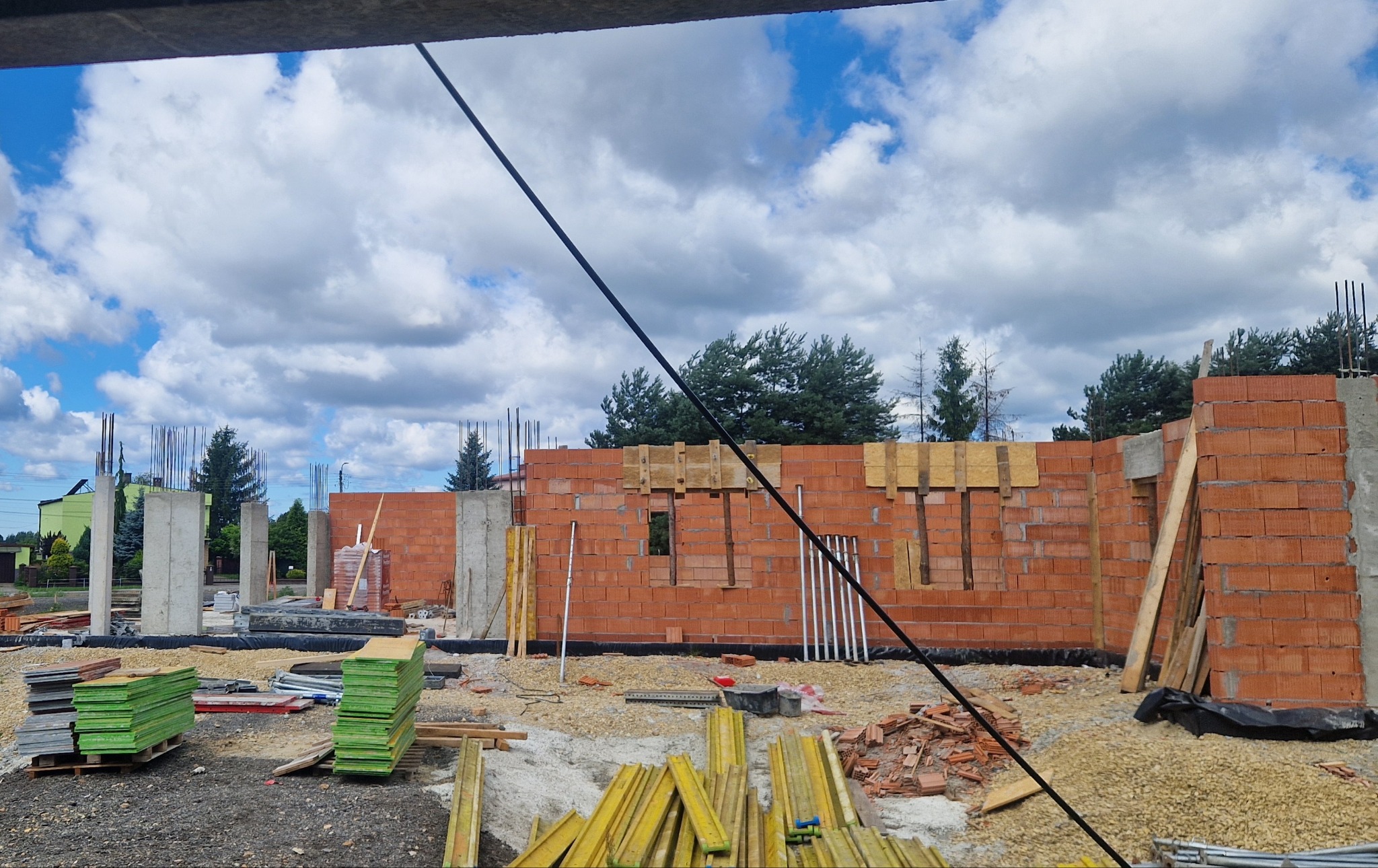 Wznoszenie ścian z czerwonej cegły na budowie domu. Widoczne betonowe słupy, zbrojenia i drewniane podpory. Niebieskie niebo z obłokami w tle.