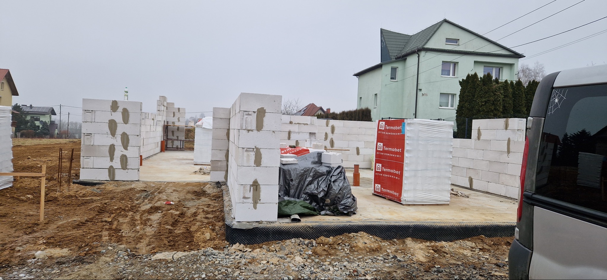 Budowa domu w toku: białe bloczki Termobet tworzą ściany na fundamencie. W tle zielony budynek i fragment samochodu dostawczego z naklejką pająka.
