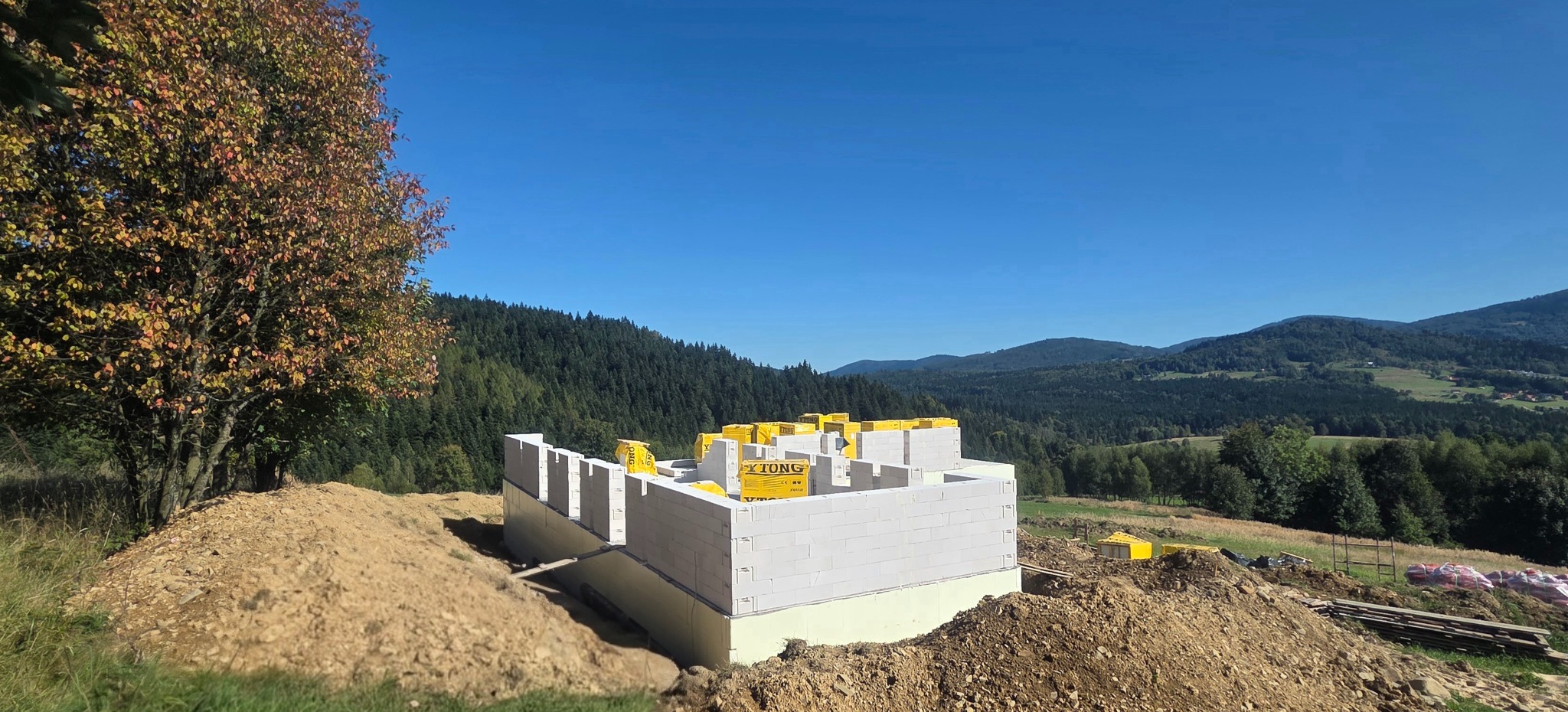 Wznoszenie ścian parteru z bloczków Ytong na fundamencie z izolacją, w tle malowniczy krajobraz górski i jesienne drzewo. Widok na plac budowy w słoneczny dzień.