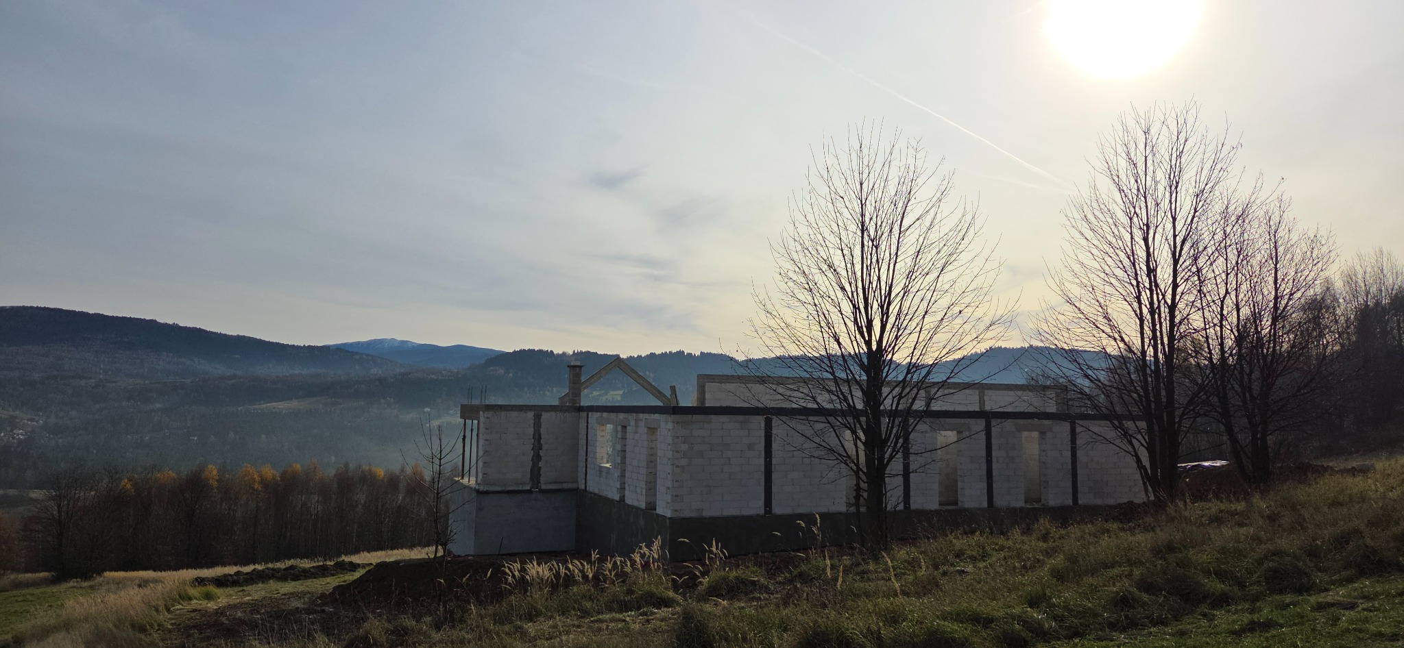 Surowy stan domu z betonu komórkowego w jesiennym krajobrazie górskim. Widoczne ściany, fundamenty i elementy konstrukcji dachu. Słońce w tle.