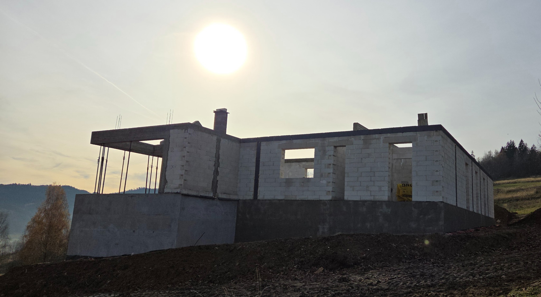 Surowy stan domu z betonu komórkowego w słoneczny dzień. Widoczne fundamenty i ściany bez tynków, na tle pagórkowatego krajobrazu. Częściowo widoczne zbrojenia.