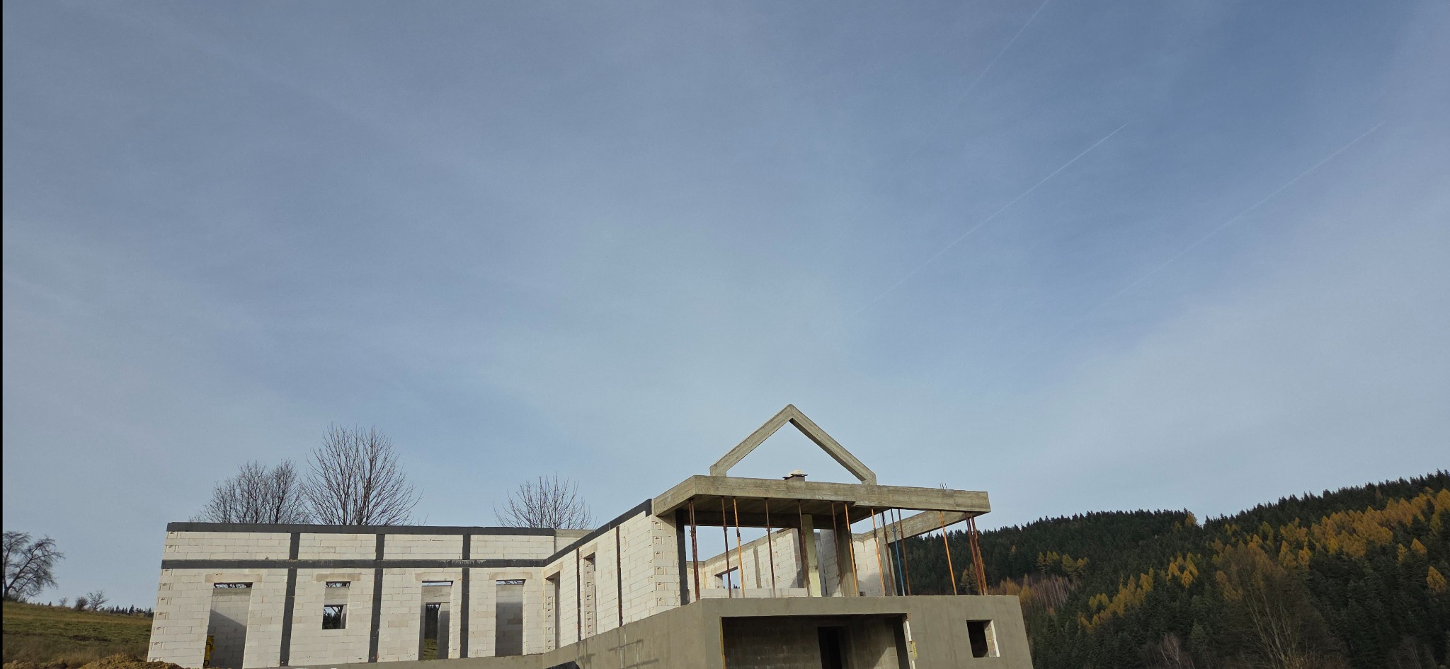 Surowy stan domu murowanego z betonu komórkowego, widoczne ściany i konstrukcja dachu na tle błękitnego nieba i lasu. Wczesny etap budowy, widok z dołu.