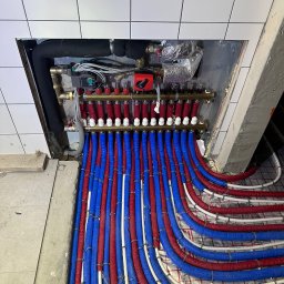 Plomero - Rozdzielacz ogrzewania podłogowego z rurami w izolacji czerwonej i niebieskiej, zamontowany w płytkiej wnęce ściennej z białymi płytkami, widok z góry.