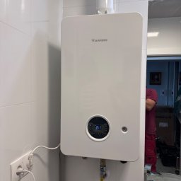 Plomero - Biały, wiszący piec gazowy Junkers na tle białych płytek. Widoczne podłączenie do gniazdka i rury. W odbiciu lustra fragment sylwetki osoby w czerwonym ubraniu.