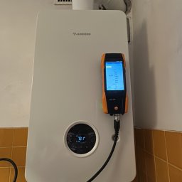 Plomero - Biały piec gazowy Junkers z podłączonym analizatorem spalin Testo 300 podczas pomiaru, widoczne rury i instalacja. Tło: białe ściany i brązowe płytki.