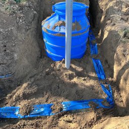 Plomero - Niebieski zbiornik w wykopie, zabezpieczony taśmą ostrzegawczą. Widoczny fragment rury pionowej. Etap instalacji w ziemi, zarys zielonej trawy w tle.