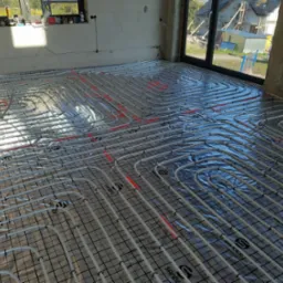 Rozłożona instalacja ogrzewania podłogowego na folii izolacyjnej w surowym wnętrzu. Widoczne rury w regularnych odstępach, przygotowane do zalania posadzką.