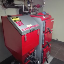 Plomero - Czerwono-szary piec Defro na paliwo stałe w kotłowni, z widoczną instalacją hydrauliczną i tabliczką informacyjną o suchym opale.