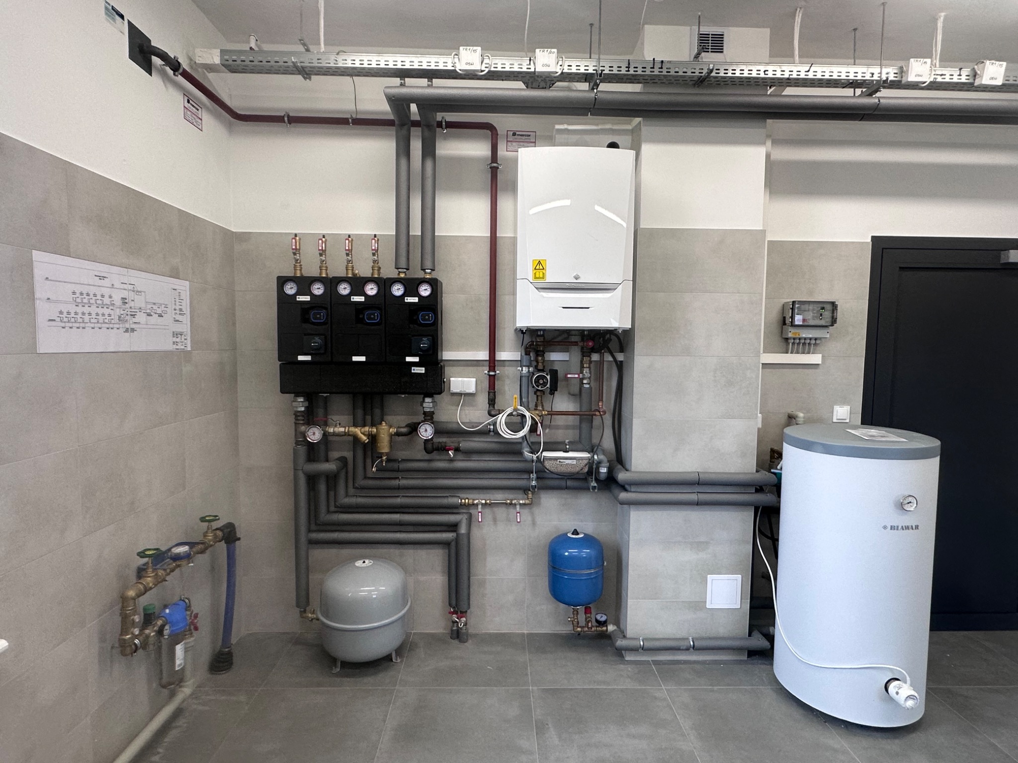 Zmontowana instalacja grzewcza z piecem gazowym, zbiornikiem i rozdzielaczami, z widocznym schematem instalacji na ścianie, w pomieszczeniu wykończonym szarą płytką.