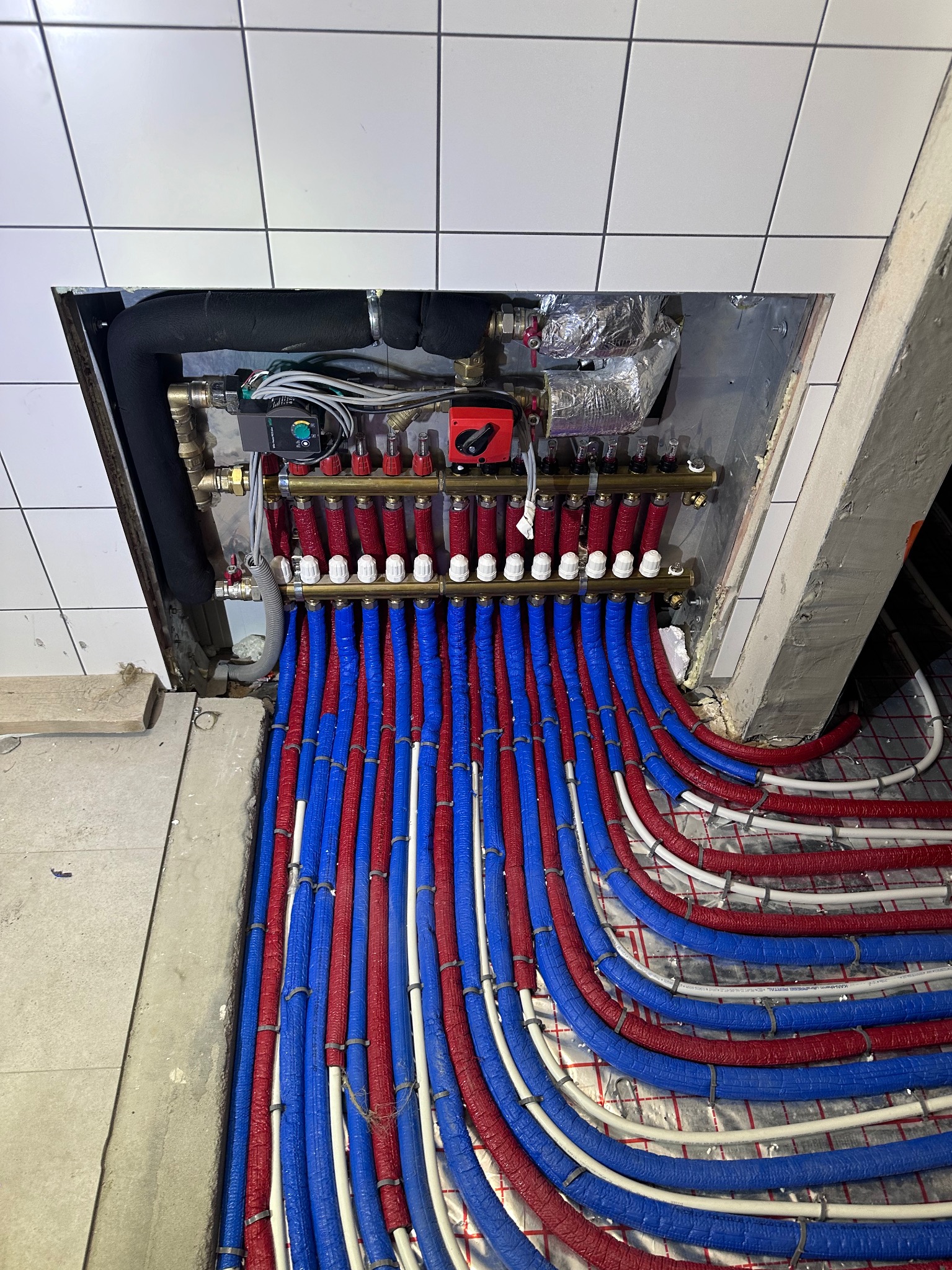 Rozdzielacz ogrzewania podłogowego z rurami w izolacji czerwonej i niebieskiej, zamontowany w płytkiej wnęce ściennej z białymi płytkami, widok z góry.