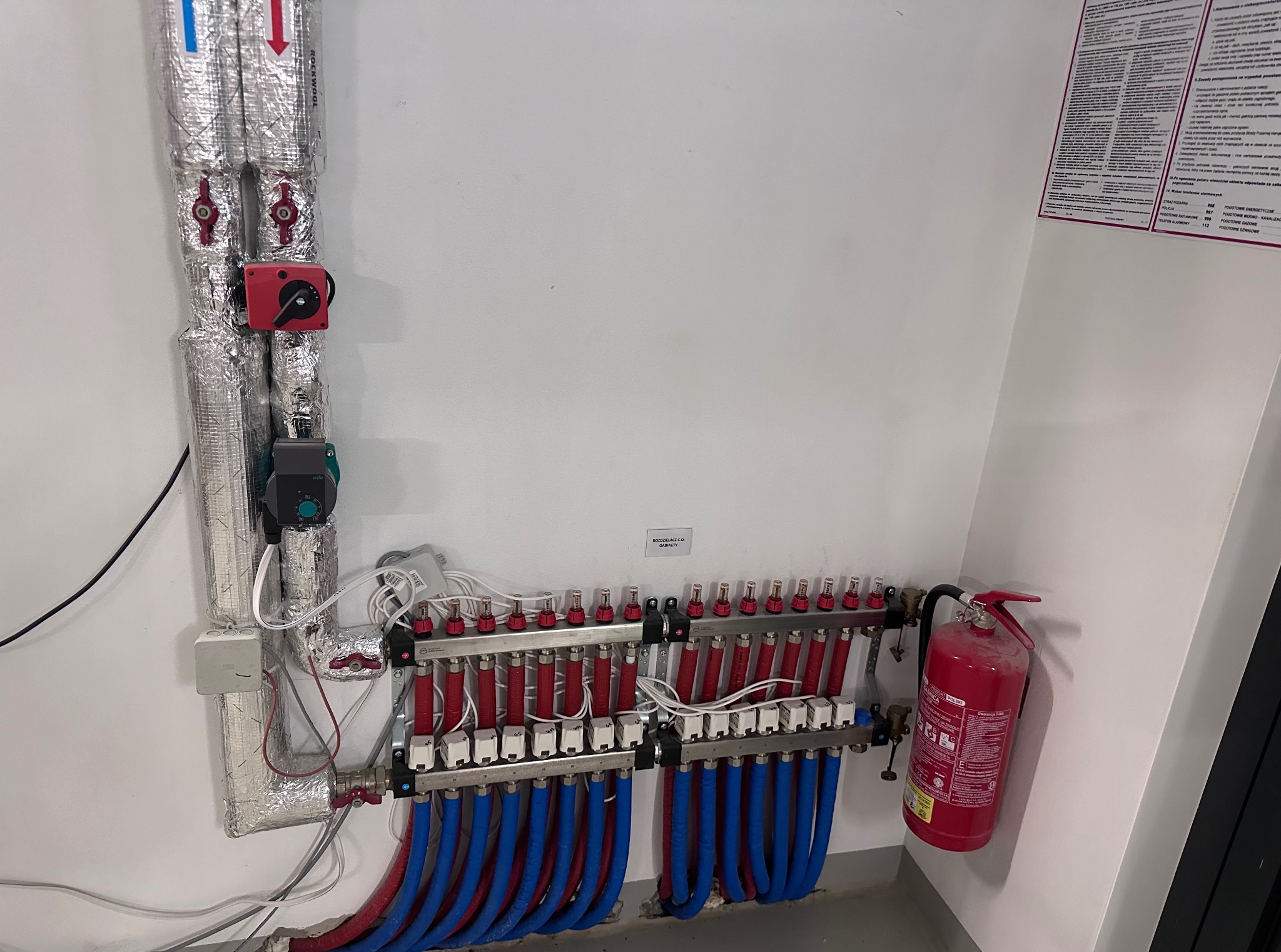 Rozdzielacz C.O. z rurami (czerwone i niebieskie) na ścianie, obok gaśnica. Instalacja w pomieszczeniu z dokumentacją techniczną na ścianie.