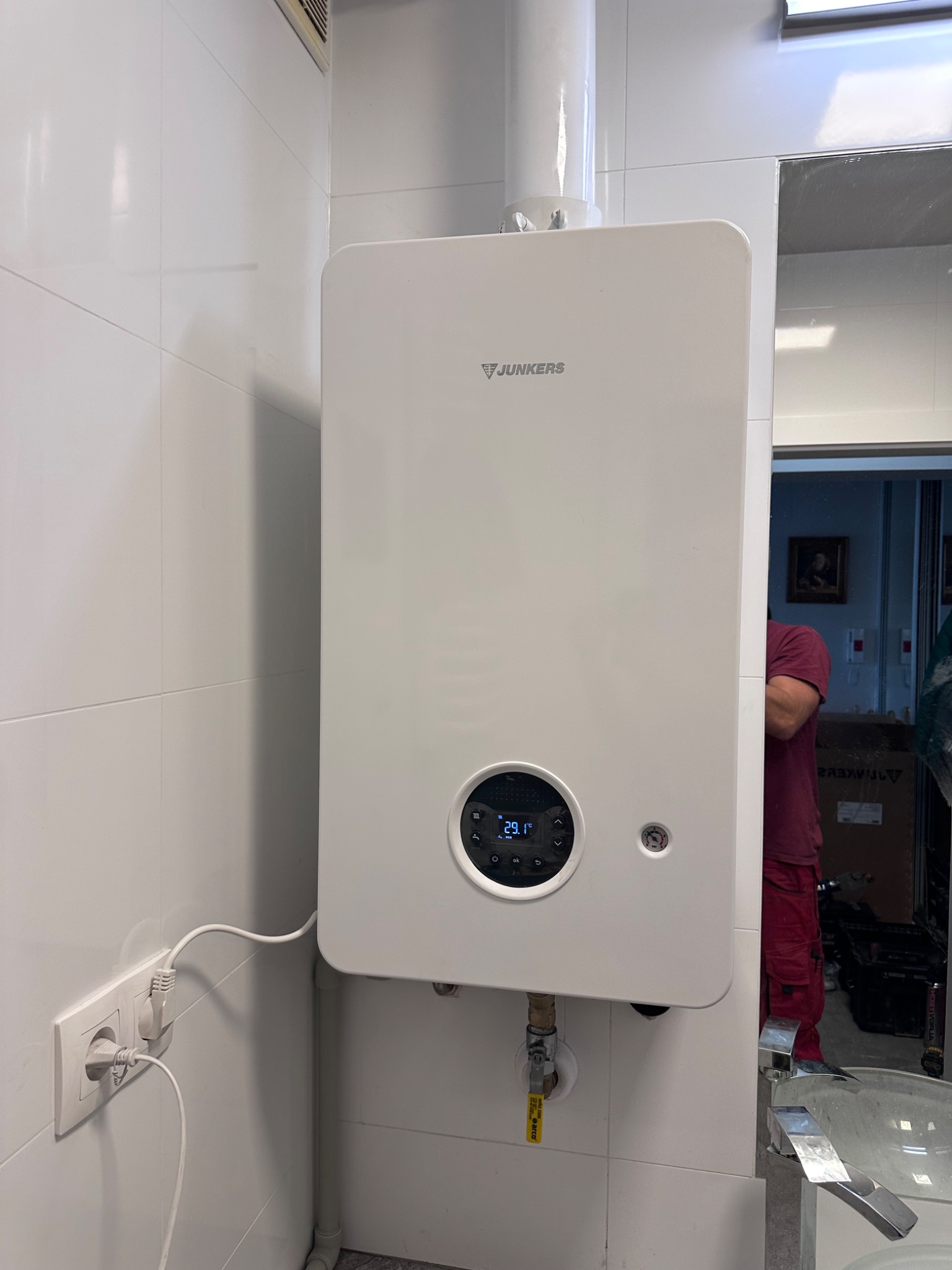 Biały, wiszący piec gazowy Junkers na tle białych płytek. Widoczne podłączenie do gniazdka i rury. W odbiciu lustra fragment sylwetki osoby w czerwonym ubraniu.