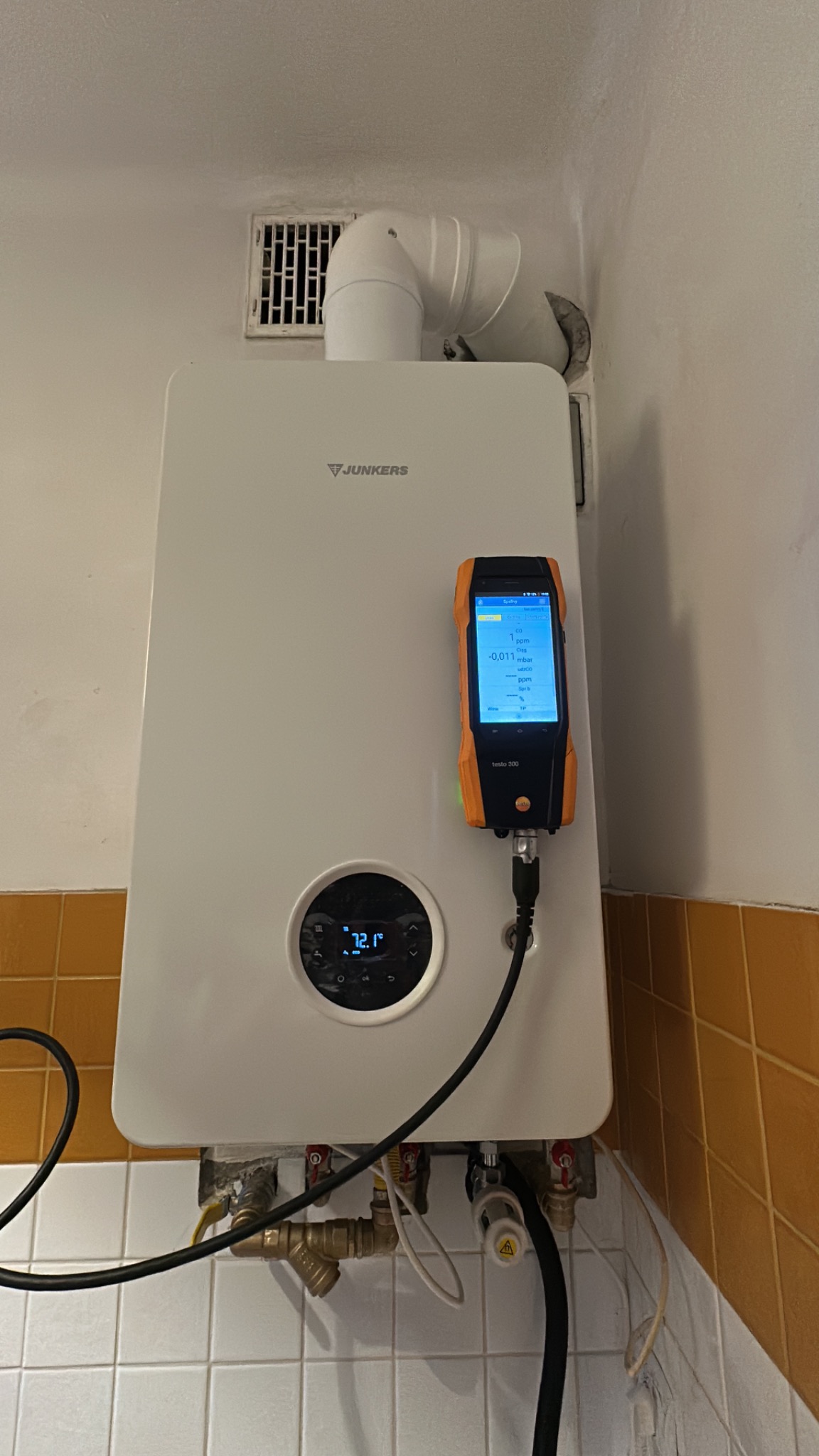 Biały piec gazowy Junkers z podłączonym analizatorem spalin Testo 300 podczas pomiaru, widoczne rury i instalacja. Tło: białe ściany i brązowe płytki.
