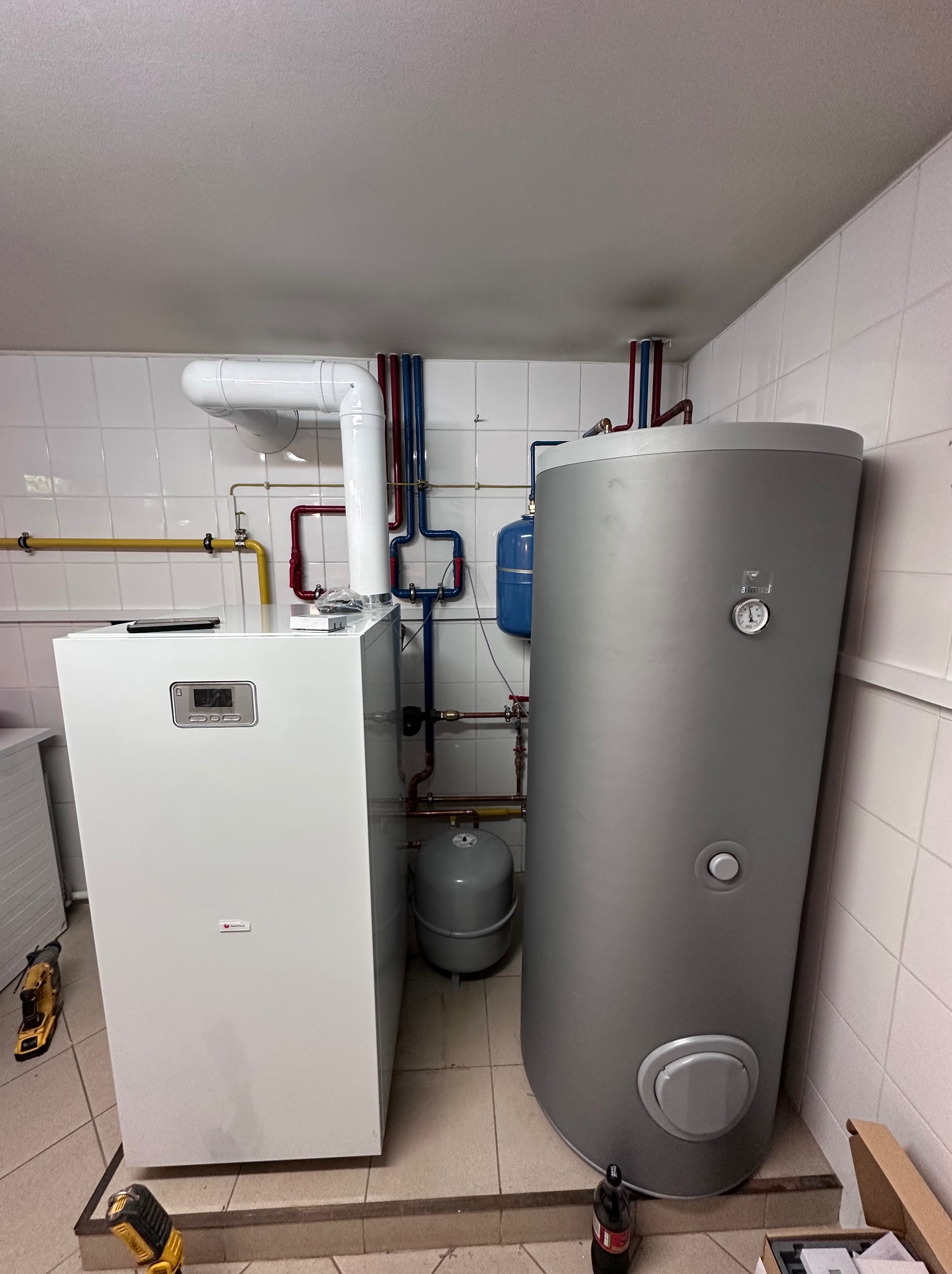 Biały piec gazowy kondensacyjny z szarym zasobnikiem ciepłej wody, rury w kolorach czerwonym i niebieskim, na podłodze narzędzia i karton.