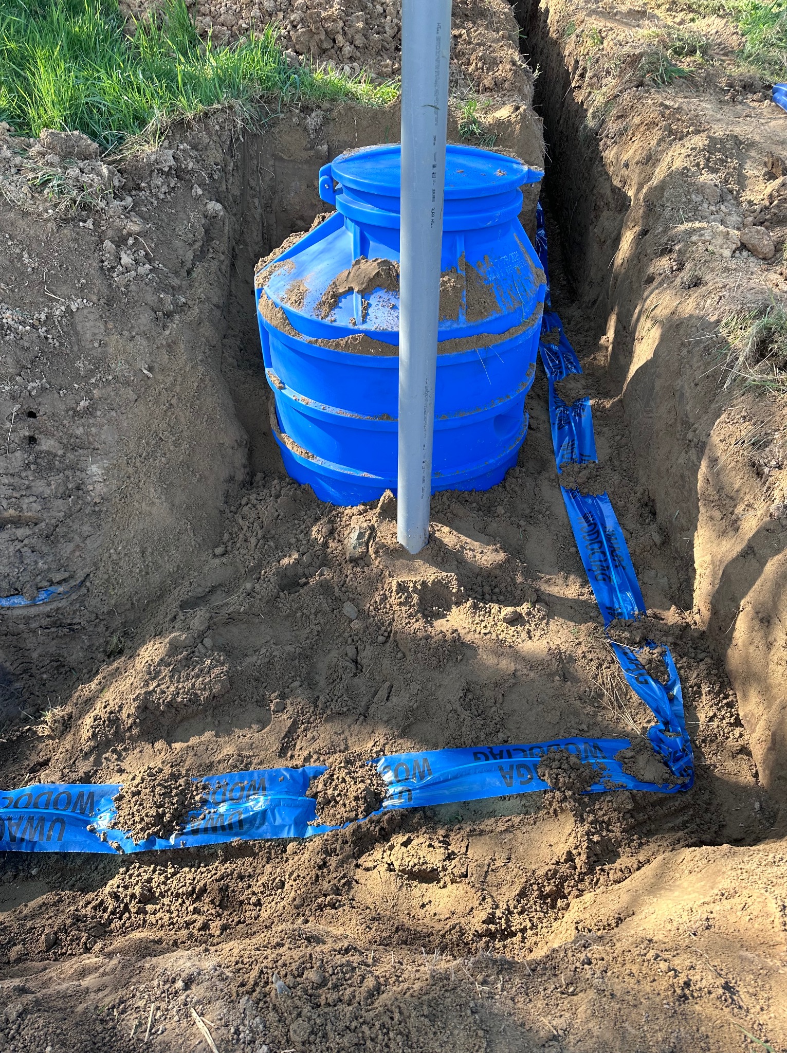 Niebieski zbiornik w wykopie, zabezpieczony taśmą ostrzegawczą. Widoczny fragment rury pionowej. Etap instalacji w ziemi, zarys zielonej trawy w tle.