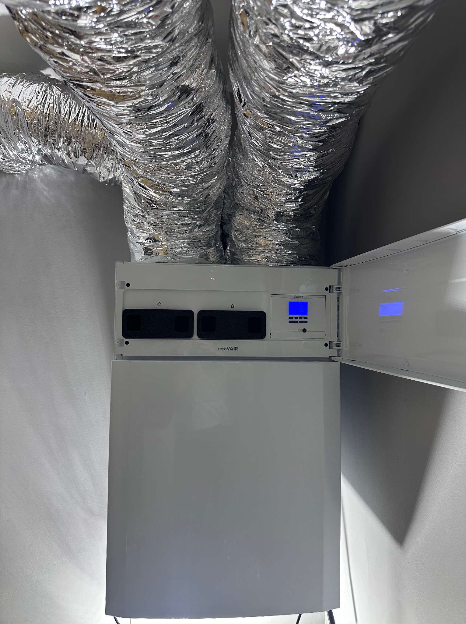 Centrala wentylacyjna z rekuperacją Vaillant recoVAIR z otwartym panelem sterowania i podłączonymi przewodami wentylacyjnymi.