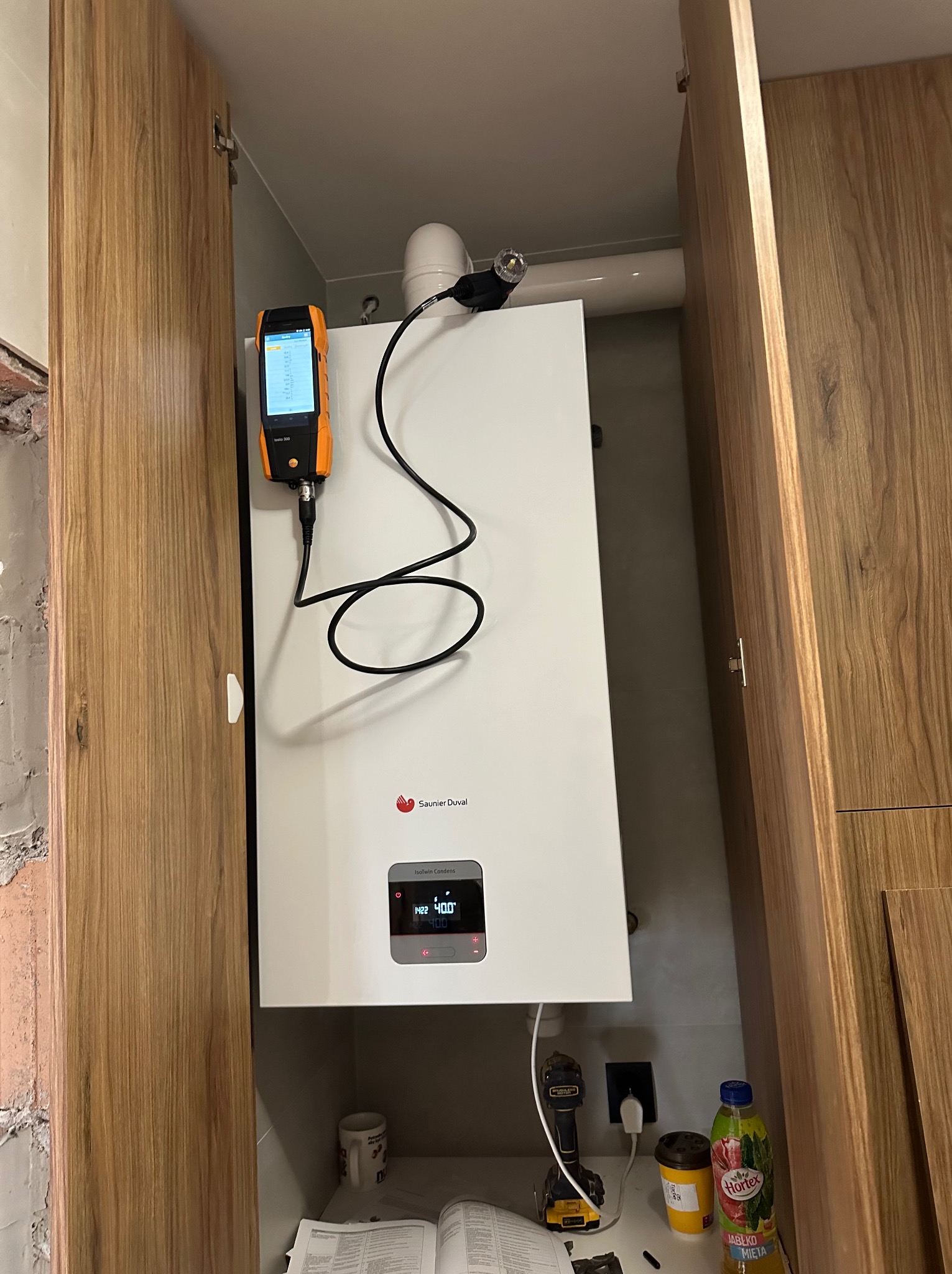 Biały piec gazowy Saunier Duval zamontowany w szafce, podłączony do analizatora spalin Testo 300. Widoczny fragment instalacji i otwarte drzwi szafki.