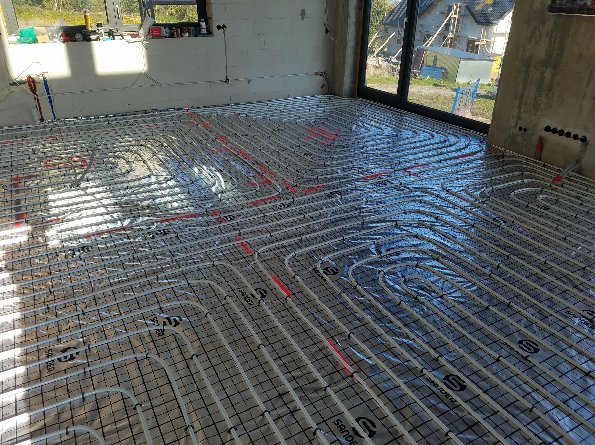 Rozłożona instalacja ogrzewania podłogowego na folii izolacyjnej w surowym wnętrzu. Widoczne rury w regularnych odstępach, przygotowane do zalania posadzką.