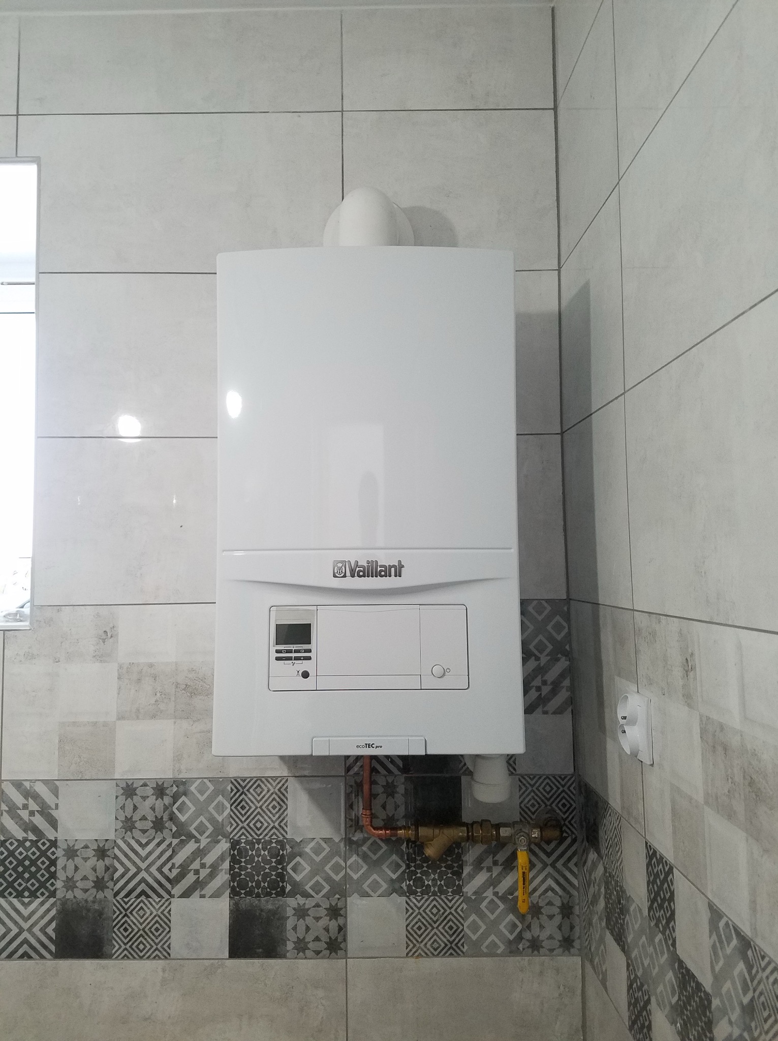 Biały, wiszący piec gazowy Vaillant na ścianie z szarymi, wzorzystymi płytkami. Widoczne rury miedziane i zawór gazowy. Górny komin.