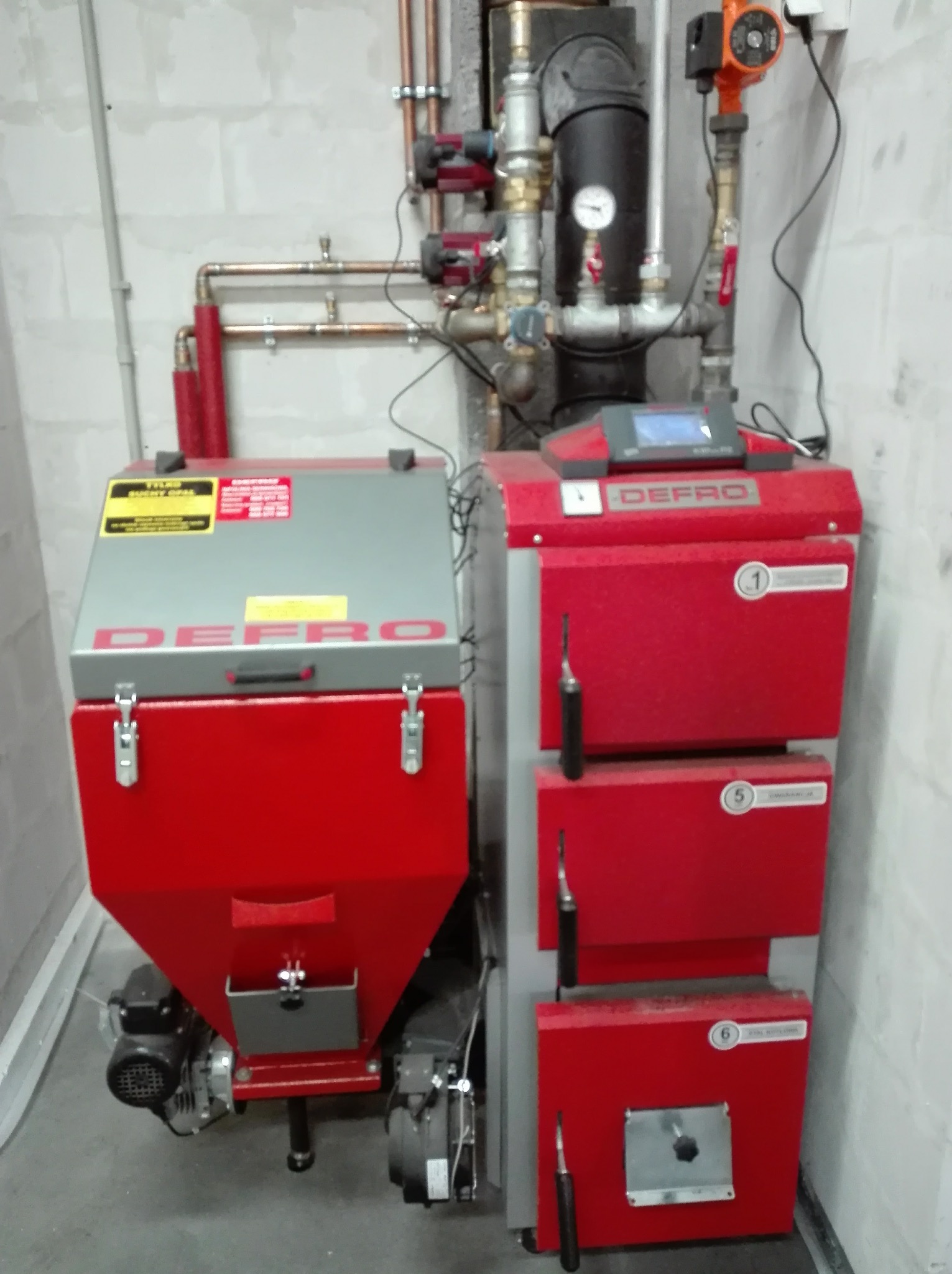 Nowoczesny, czerwono-szary piec Defro z podajnikiem, zainstalowany w kotłowni. Widoczne rury instalacji grzewczej i elementy sterujące.