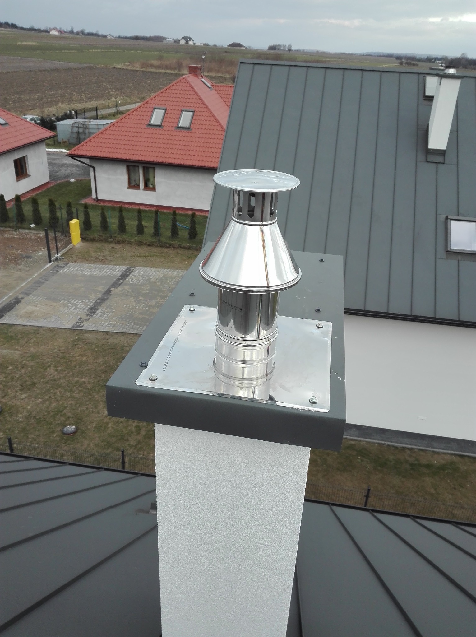 Błyszczący, metalowy komin na dachu z blachy trapezowej, zamontowany na białym kominie. W tle budynki mieszkalne i pole uprawne.