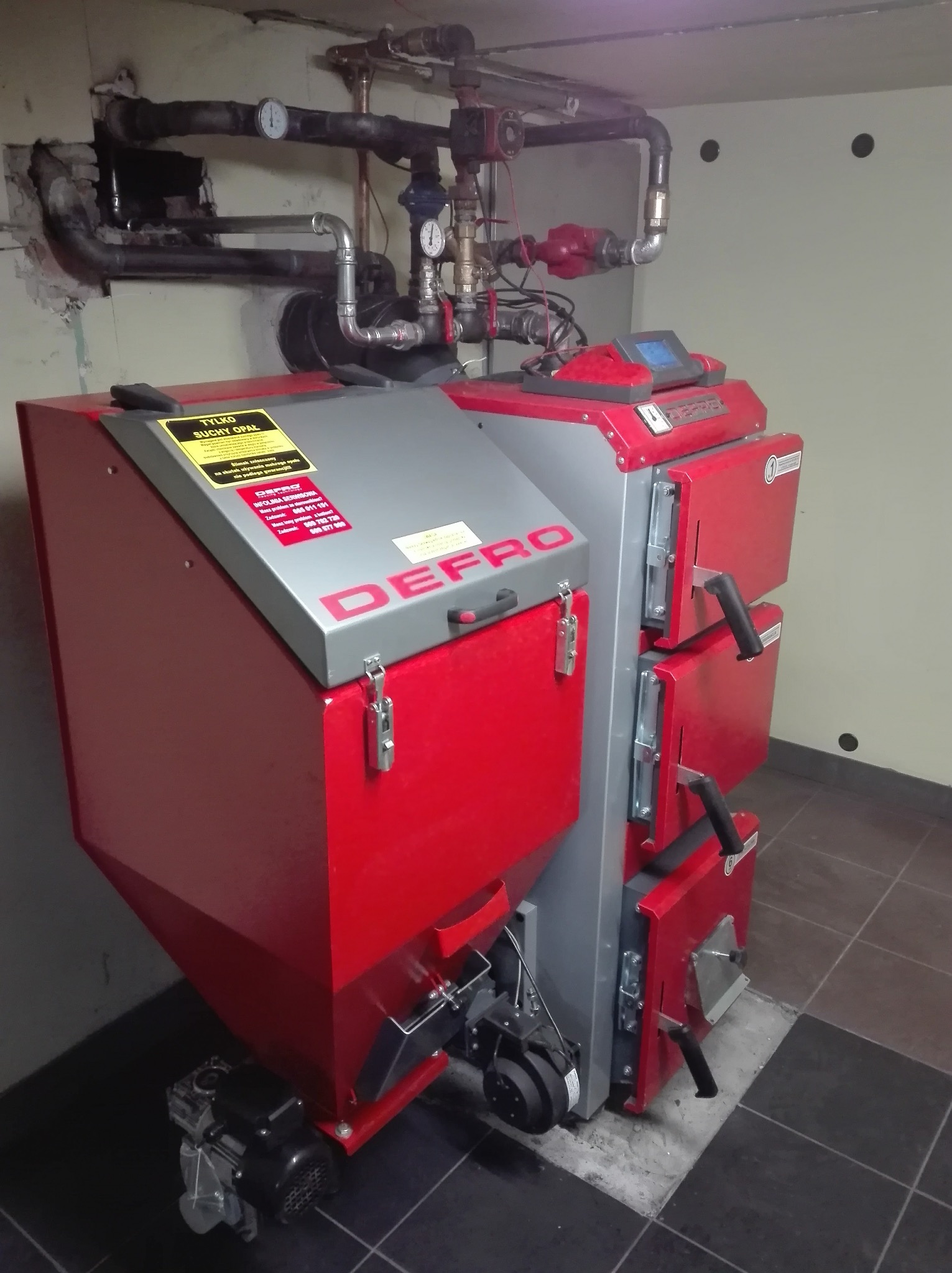 Czerwono-szary piec Defro na paliwo stałe w kotłowni, z widoczną instalacją hydrauliczną i tabliczką informacyjną o suchym opale.