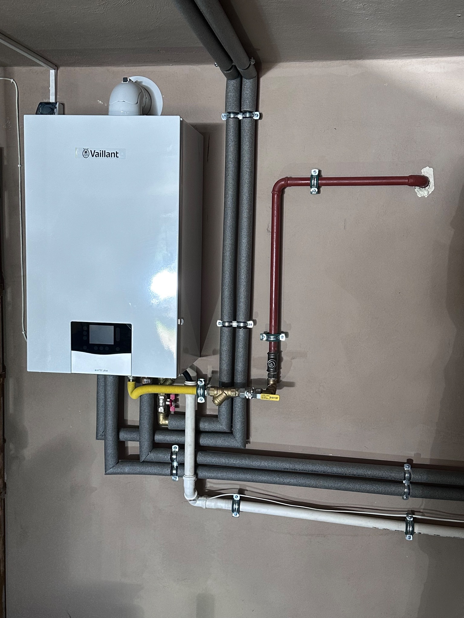 Biały piec gazowy Vaillant, zamontowany na ścianie, z podłączonymi rurami w izolacji, rura gazowa żółta. Instalacja grzewcza w piwnicy.