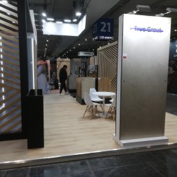 Janusz Jasiński - Stoisko targowe z panelami podłogowymi i ścianami z drewnianymi lamelami. Widoczny stolik z krzesłami i osoba w tle. Oświetlenie sufitowe.