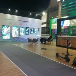 Janusz Jasiński - Stoisko targowe z panelami LCD o wysokiej jasności, stolikami i krzesłami dla odwiedzających oraz skuterem elektrycznym. Widoczny bar z menu kawowym.