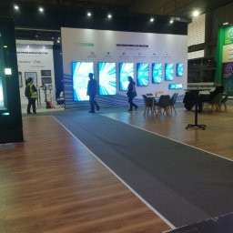 Janusz Jasiński - Stoisko targowe z panelami LED Dynascan prezentujące jasne ekrany LCD. Osoby przechodzą obok, a w tle widoczne są elementy konstrukcyjne hali.