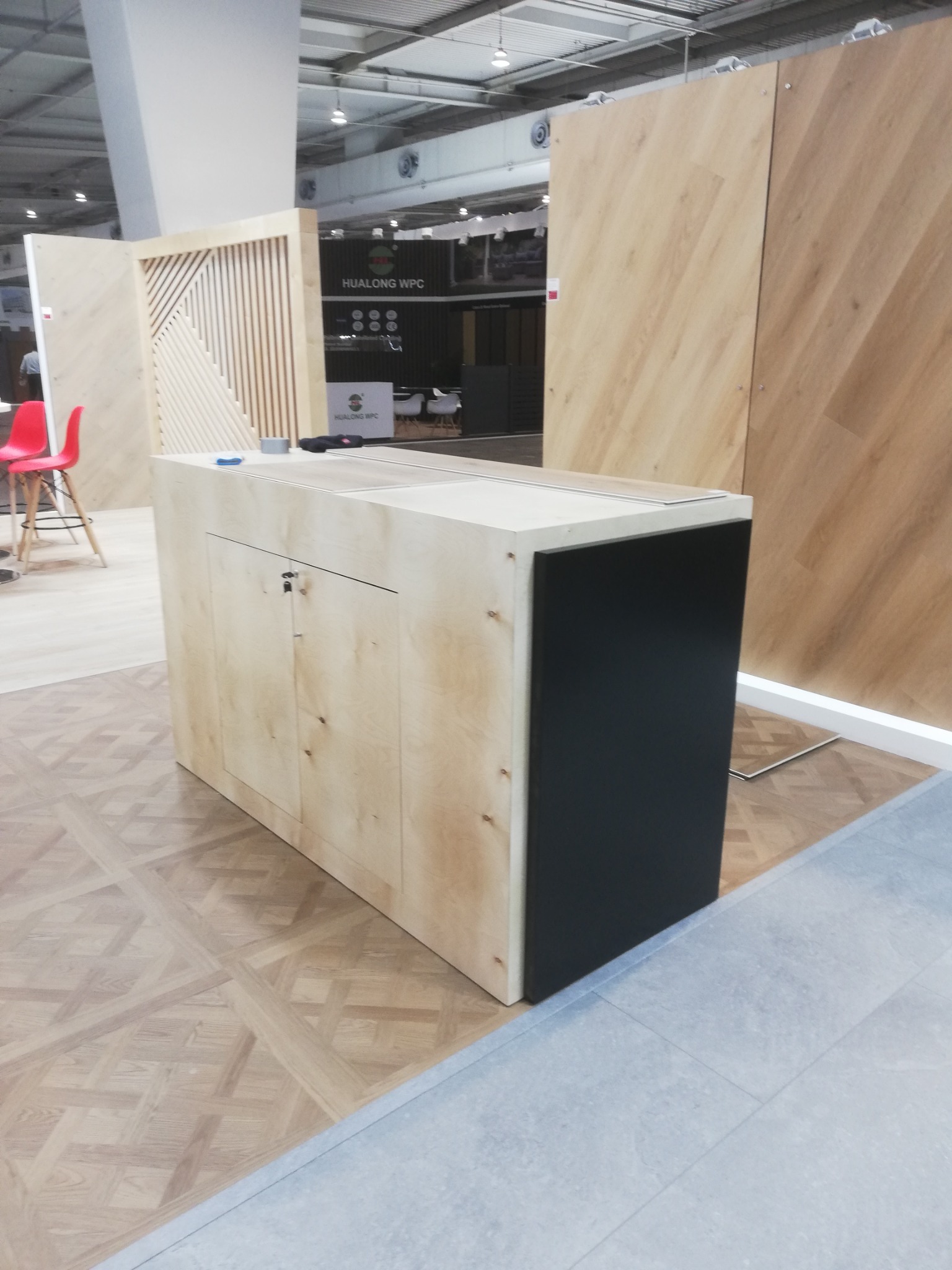 Drewniana zabudowa meblowa z czarnym bokiem na tle paneli podłogowych i ścianek działowych na targach. Widoczne zamki w szafkach i fragment podłogi w jodełkę.