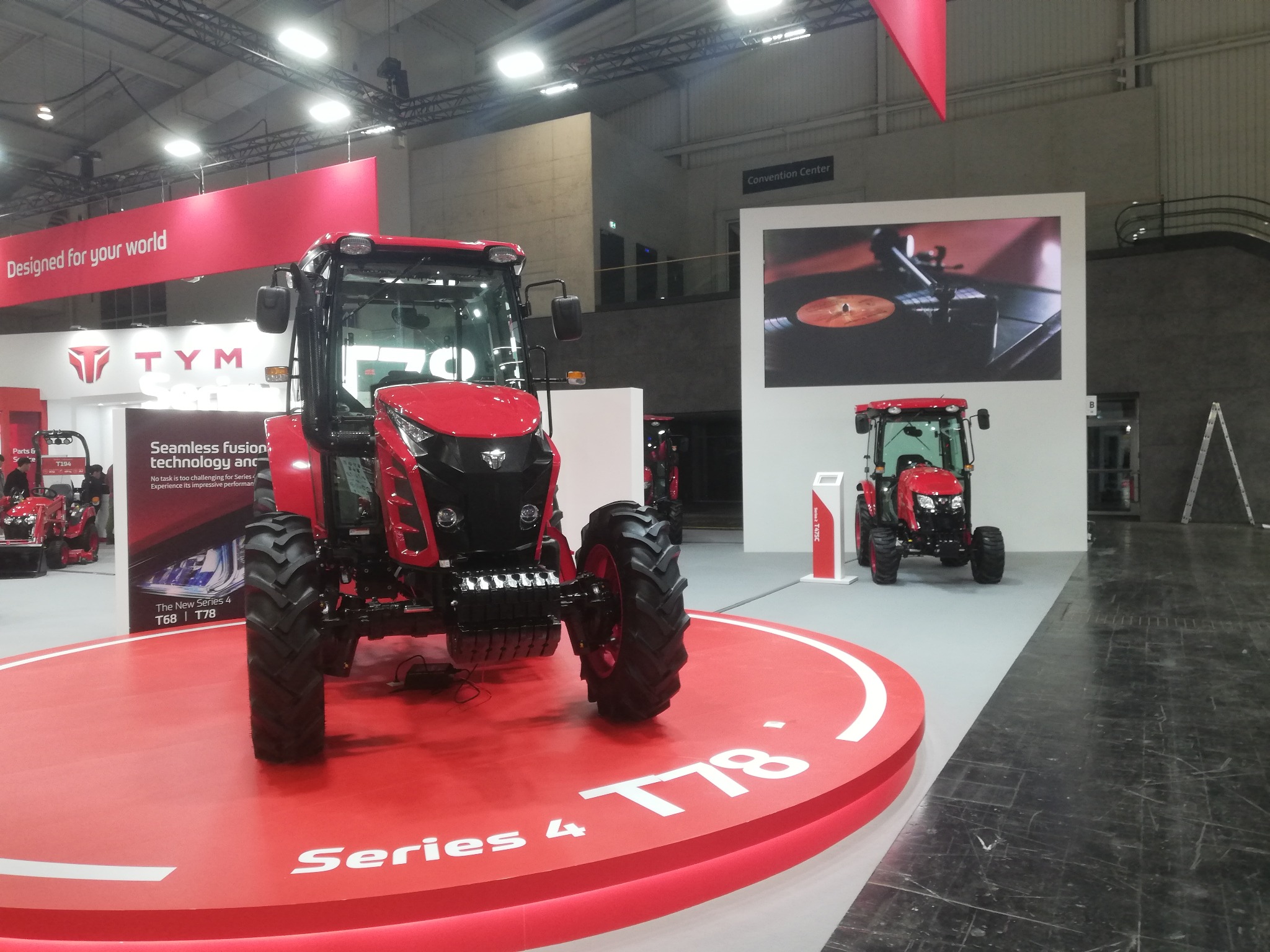 Czerwony ciągnik TYM Series 4 T78 na czerwonym podeście z napisem, prezentowany na targach w hali Convention Center. W tle drugi ciągnik i reklamy.