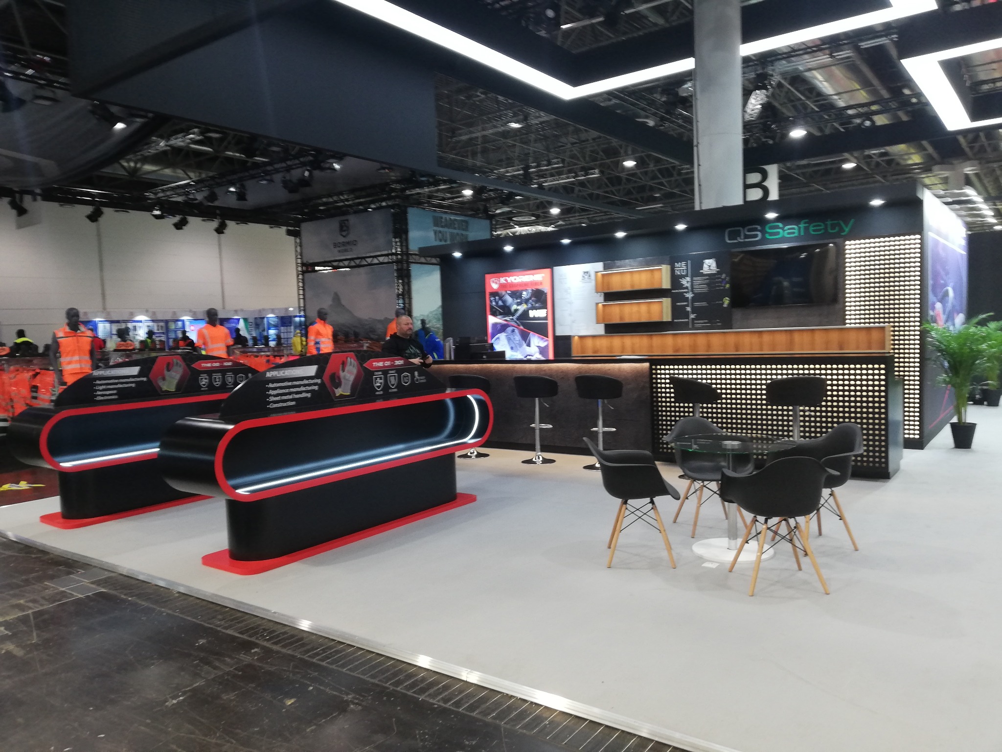 Nowoczesne stoisko targowe QS Safety z ladą, stołkami barowymi i stolikiem z krzesłami. Widoczne elementy graficzne i oświetlenie. Przestrzeń wystawiennicza.