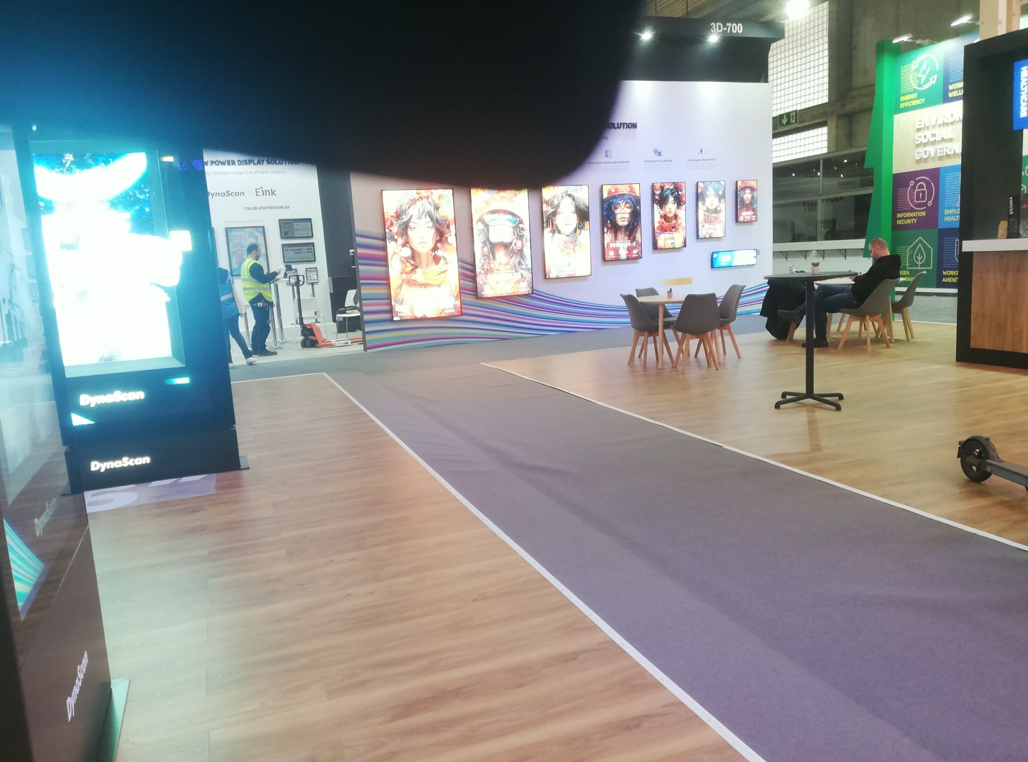 Wnętrze stoiska targowego z panelami LED z grafikami, stolikami i osobą siedzącą. Podłoga drewniana, szara wykładzina. Widoczne logo DynaScan.