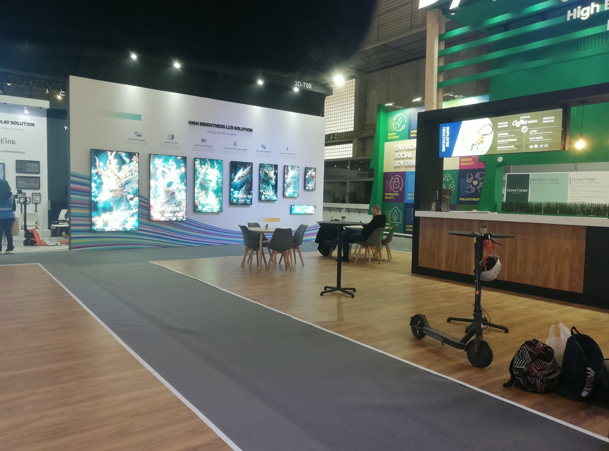 Stoisko targowe z panelami LCD o wysokiej jasności, stolikami i krzesłami dla odwiedzających oraz skuterem elektrycznym. Widoczny bar z menu kawowym.