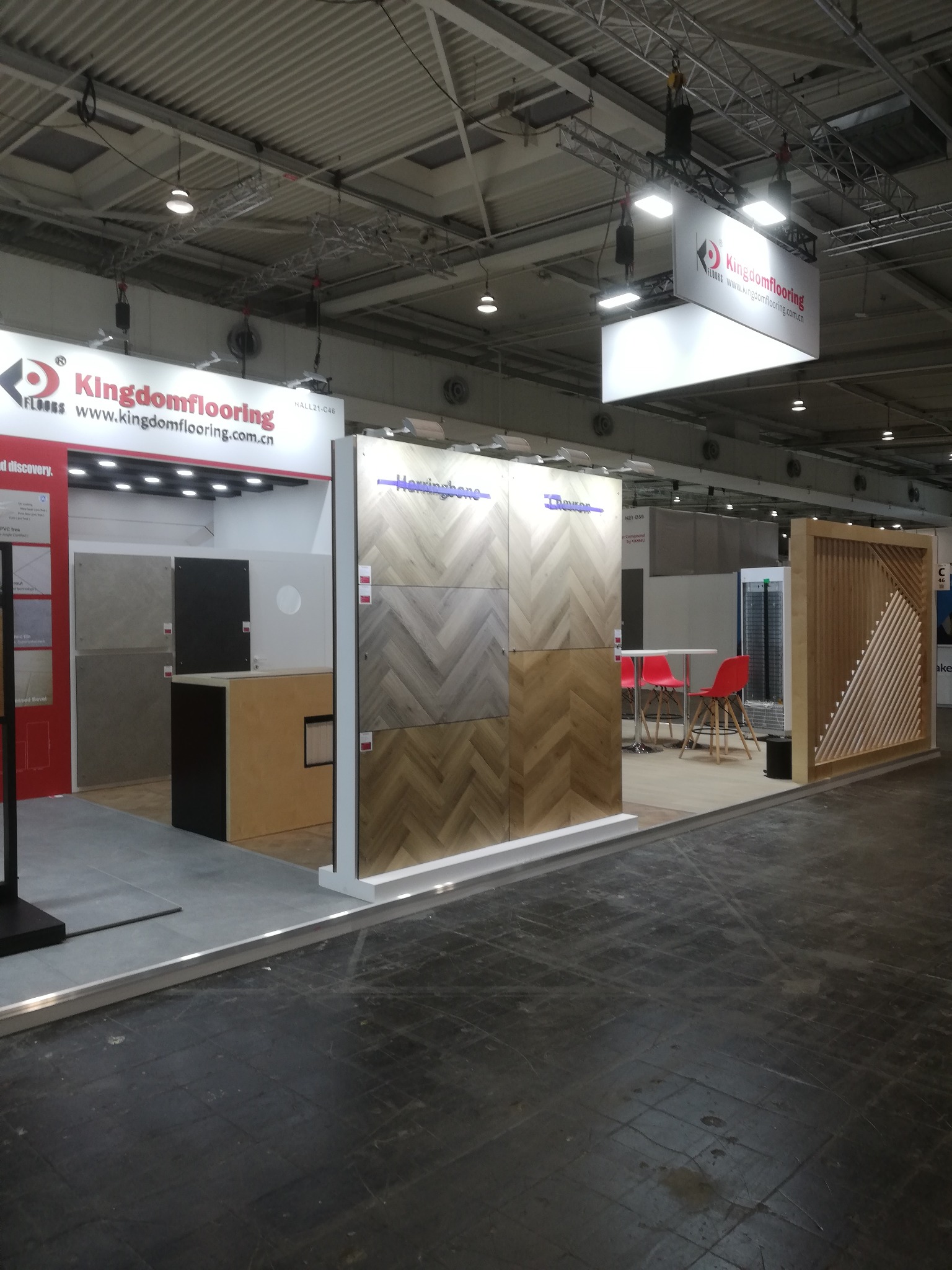 Ekspozycja paneli podłogowych w jodełkę na stoisku targowym Kingdomflooring, z drewnianą ścianką działową i czerwonymi krzesłami w tle. Oświetlenie sufitowe.