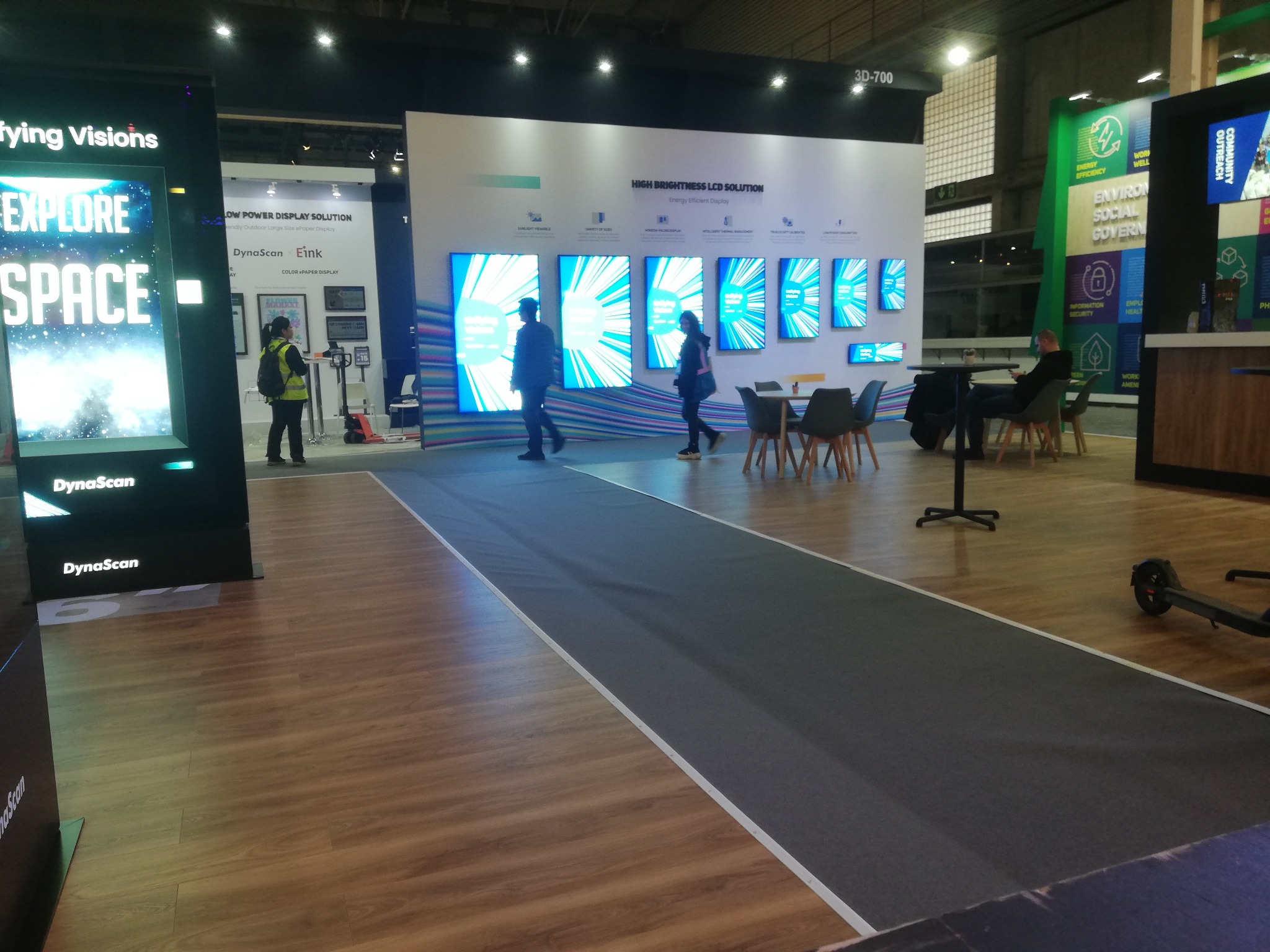 Stoisko targowe z panelami LED Dynascan prezentujące jasne ekrany LCD. Osoby przechodzą obok, a w tle widoczne są elementy konstrukcyjne hali.